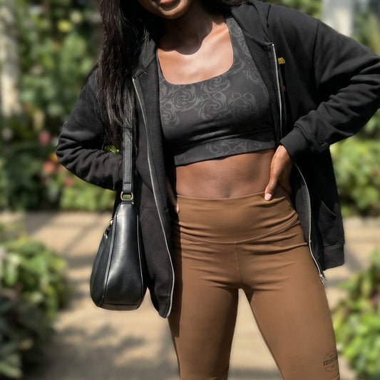 a black woman wearing brown biker shorts a black sports bra(with grey roses  as prints). She wears a black zip-up hoodie on top of the sports bra.