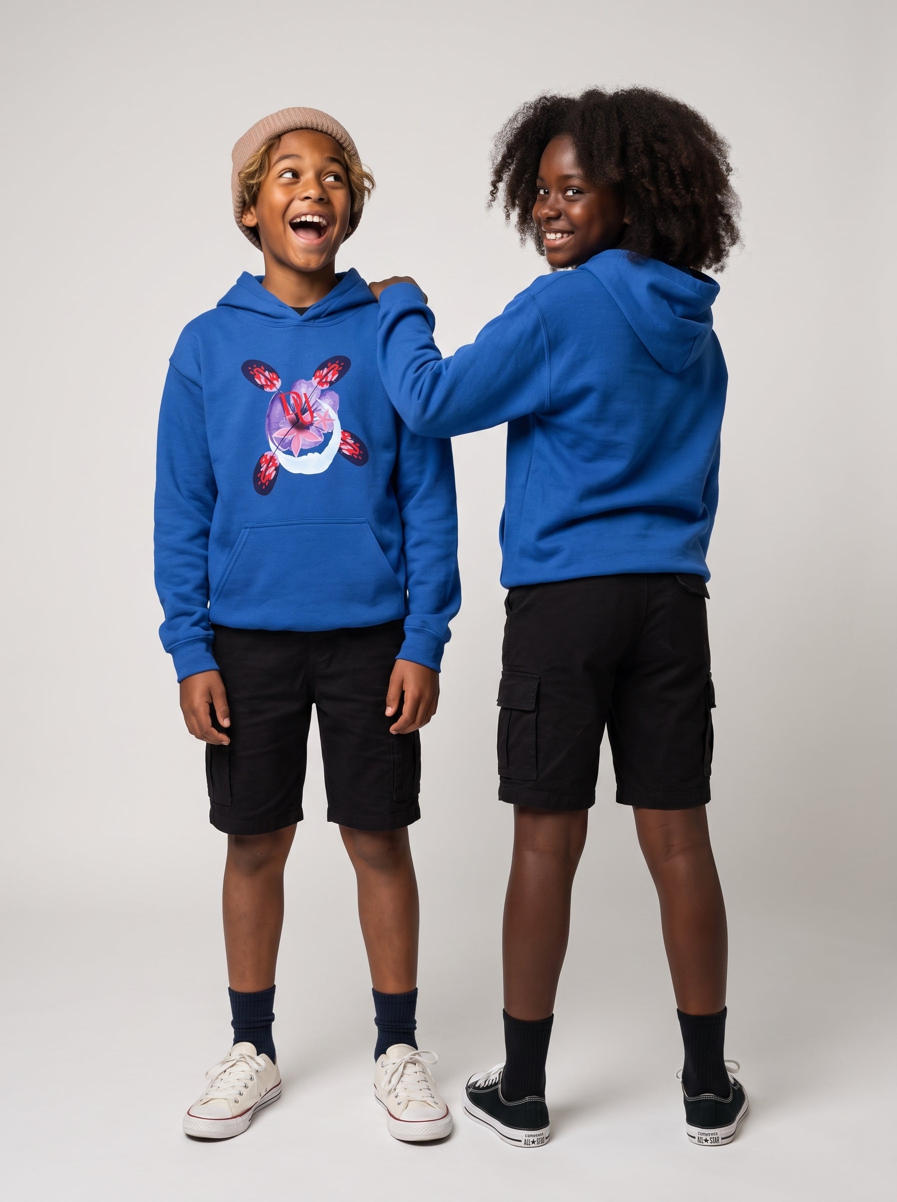 Two children modeling bright blue hoodies in a studio setting. The child on the left wears a tan beanie, black cargo shorts, dark socks, and white sneakers, while the child on the right wears black cargo shorts and black sneakers.