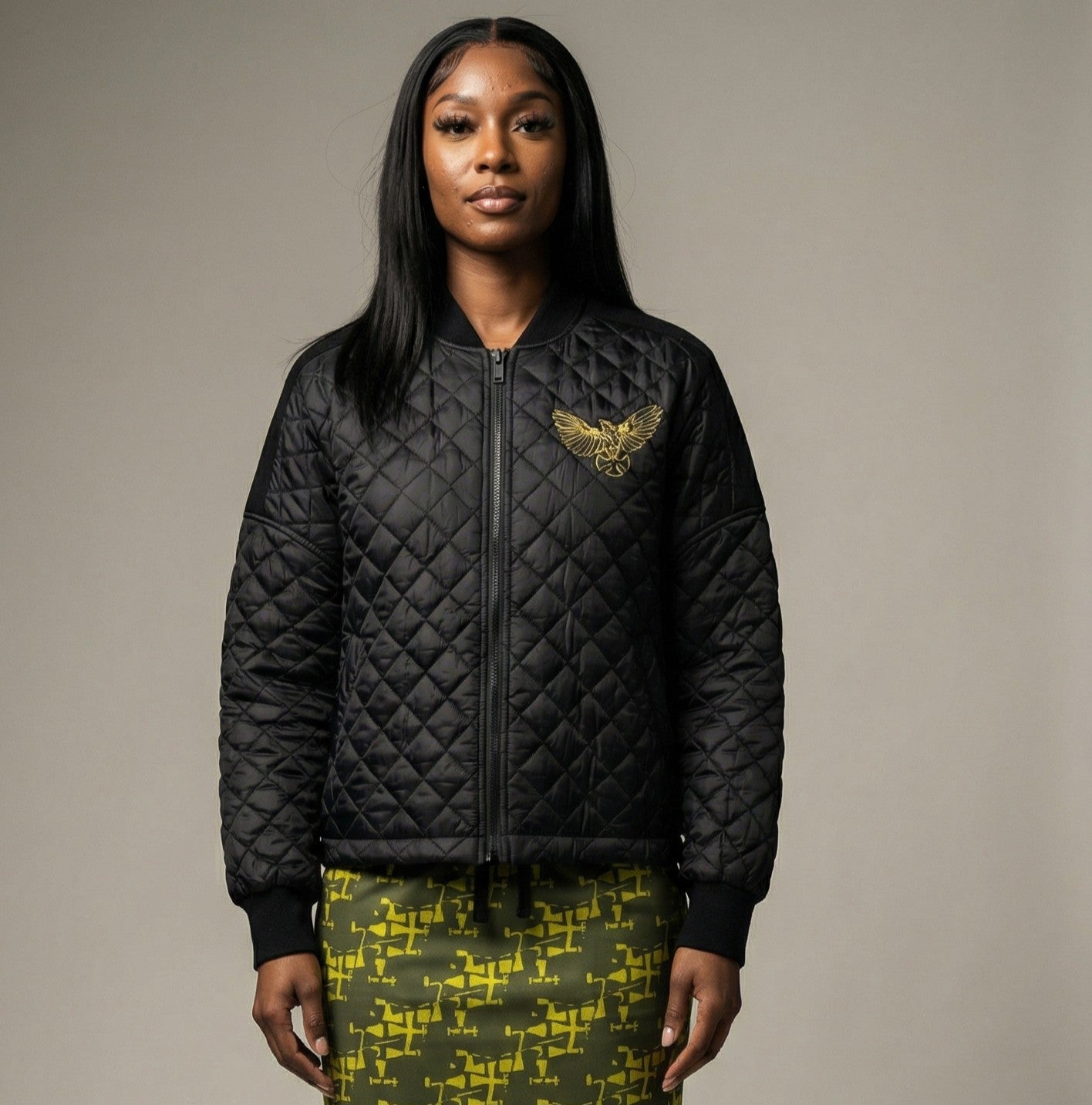 Woman wearing a black quilted jacket with a gold eagle emblem on a neutral background
