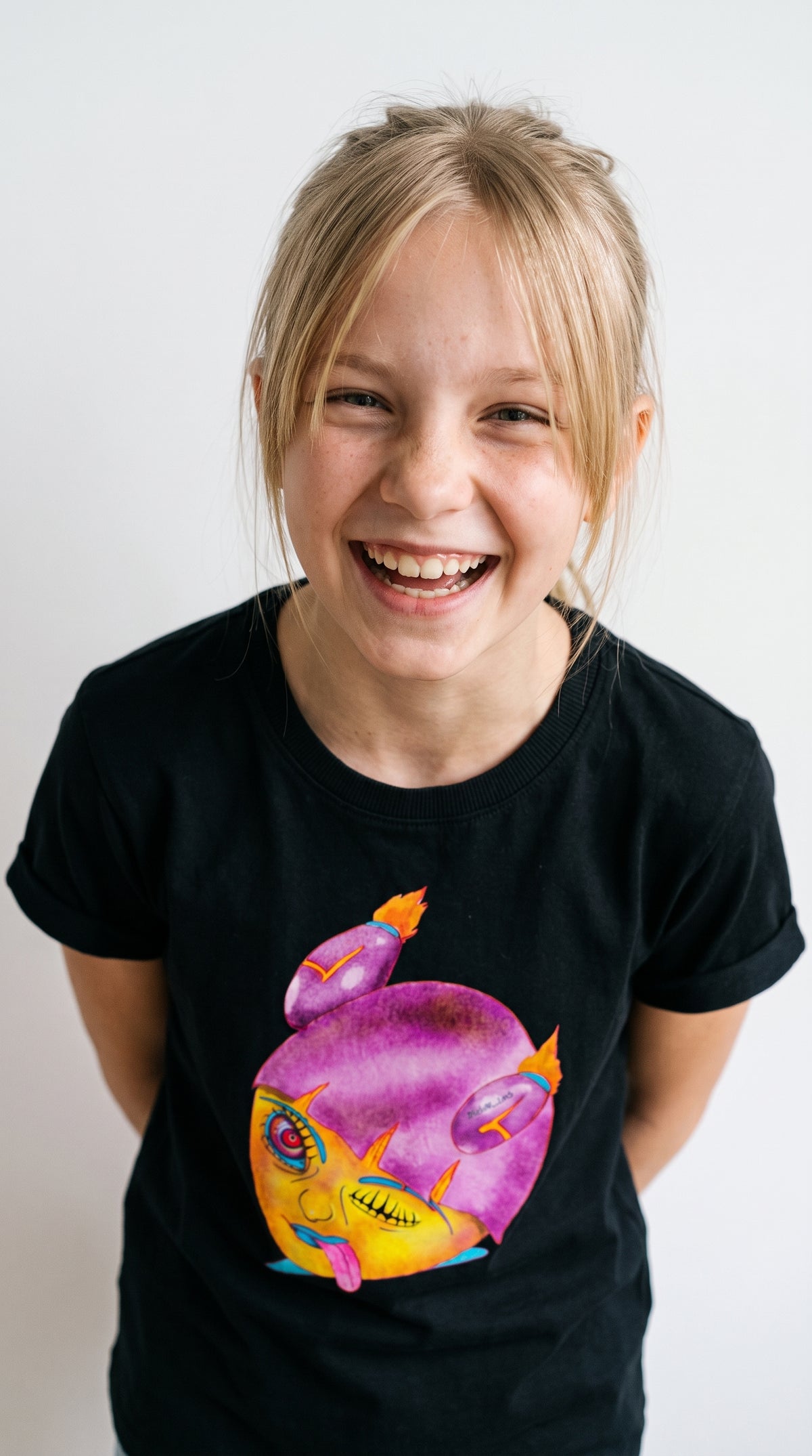 A young girl with blonde hair smiling while wearing a black t-shirt with a colorful graphic of a purple and yellow creature.